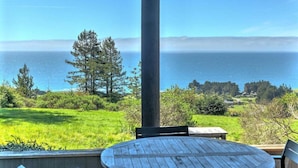 A wooden deck with a round table and chairs, overlooking a body of water and a distant shoreline with trees. - Redwood | Japanese-Inspired Retreat by the Sea (Sea Ranch)