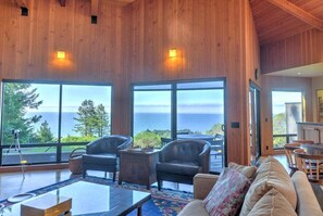 A spacious living area with large windows offering a view of the ocean, wooden paneling on the walls, and a cozy seating arrangement.