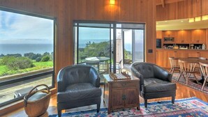 A modern interior with a large window offering a view of the sea, a dining area with wooden furniture, and a kitchen with wooden cabinets. - Redwood | Japanese-Inspired Retreat by the Sea (Sea Ranch)