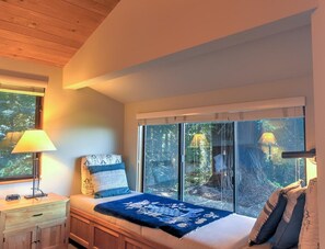Een slaapkamer met een groot raam, een bed met een blauwe deken, een nachtkastje met een lamp en een houten kast.