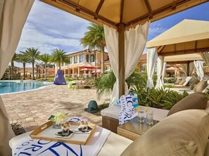 Un espace aménagé au bord de la piscine, comprenant une chaise longue, une table avec un plateau, et offrant une vue sur la piscine et les bâtiments du complexe hôtelier.
