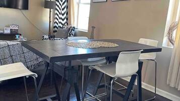 A dining area with a rectangular table, four white chairs, a black and white chevron curtain, a flat-screen TV mounted on the wall, and framed photographs on the wall.