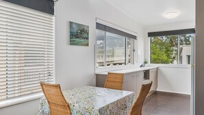 A modern kitchen with white cabinetry, a large window, and a dining area with a table and chairs. - H2O Holiday Apartments - Unit 5 (NORTH HAVEN)