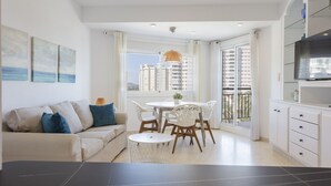 A modern living room with a sofa, dining table, and chairs, a kitchen area with cabinets, and a balcony with a view of buildings. - Cosy apartment decorated, with wifi, in front of the beach - only families (Playa de Gandía)