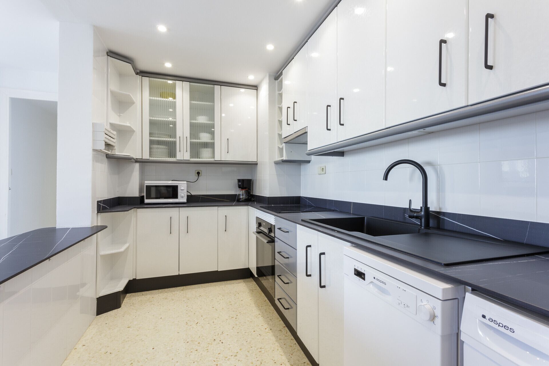 Una cocina moderna con armarios blancos, encimera negra, microondas integrado, lavavajillas y fregadero.