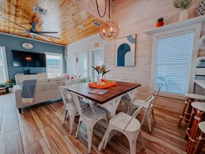 Un interior moderno con techo de madera, un comedor con sillas blancas, un televisor montado en la pared y un amplio ventanal con persianas.