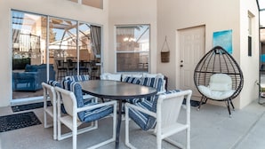 Un espace patio avec une table ronde et des chaises blanches rembourrées, un fauteuil en osier et une porte avec une pancarte.