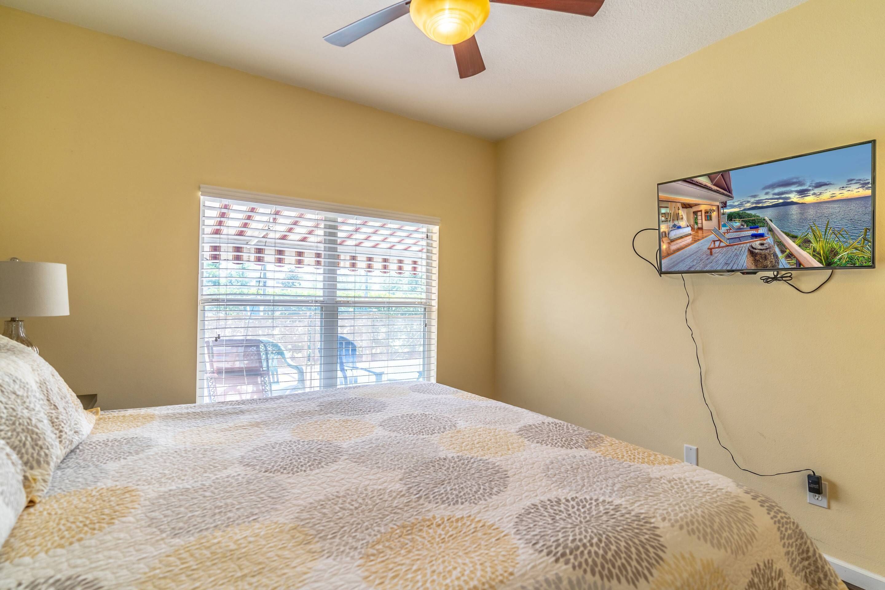 Une chambre avec un lit, un ventilateur de plafond, une fenêtre avec des stores, un téléviseur fixé au mur et une prise de courant.