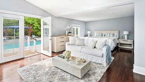 A modern bedroom with a large bed, a nightstand, a dresser, and a view of a pool through glass doors.