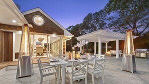 An outdoor patio with a dining table set for four, surrounded by white chairs. There are two standing heaters on either side of the table.