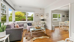 A spacious living room with a grey sofa, a glass coffee table, and a cowhide rug. There are large windows with a view of greenery outside.