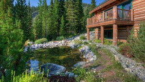 A log cabin with a balcony overlooking a pond surrounded by dense forest.