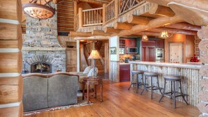 A rustic cabin interior with a stone fireplace, wooden beams, a loft bedroom, a kitchen, and a bar area with stools.