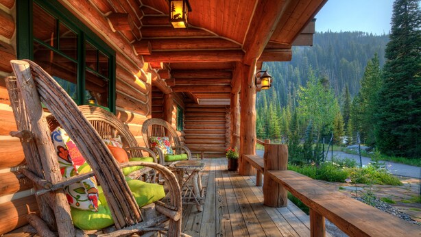 A log cabin with a wooden porch, rustic furniture, and a view of a forested valley.
