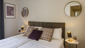 Ein ordentlich bezogenes Bett mit Kissen, ein Nachttisch mit Lampe, ein gerahmtes Bild und ein runder Spiegel an der Wand.