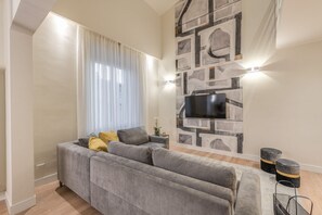 A modern living room with a grey sofa, a flat-screen TV mounted on a stone-patterned wall, and a window with sheer curtains.
