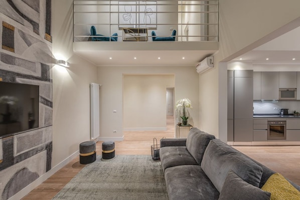 A modern living room with a grey sofa, a TV, and a staircase leading to a balcony with blue chairs.