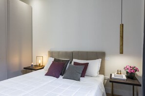 A neatly made bed with a white duvet, a headboard, and multiple pillows. There is a bedside table with a lamp and a small vase with flowers.