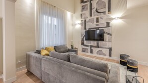 Ein modernes Wohnzimmer mit grauem Sofa, einem Flachbildfernseher an einer Wand mit Steinmusterung und einem Fenster mit durchsichtigen Vorhängen.