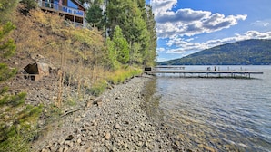 A lakeside cabin with a dock extending into the water, surrounded by dense forest and a clear sky. - Lake Pend Oreille Home w/ Dock & Paddle Boards (Sagle)