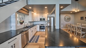 A modern kitchen with white cabinets, a black countertop, stainless steel appliances, and a dining area with wooden chairs. - Lake Pend Oreille Home w/ Dock & Paddle Boards (Sagle)