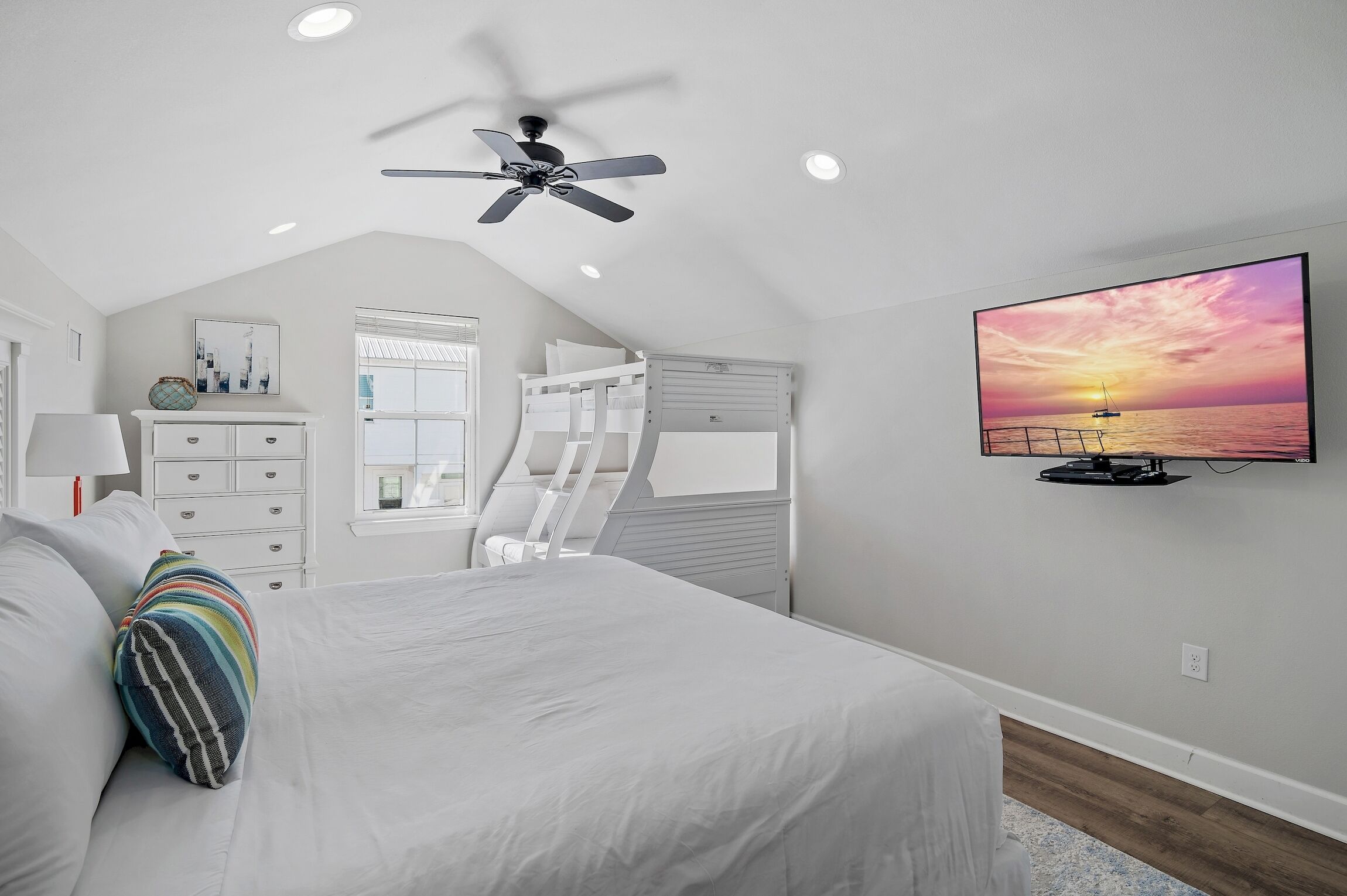A bedroom with a bed, a ceiling fan, a TV displaying a sunset, a window, and a white dresser.