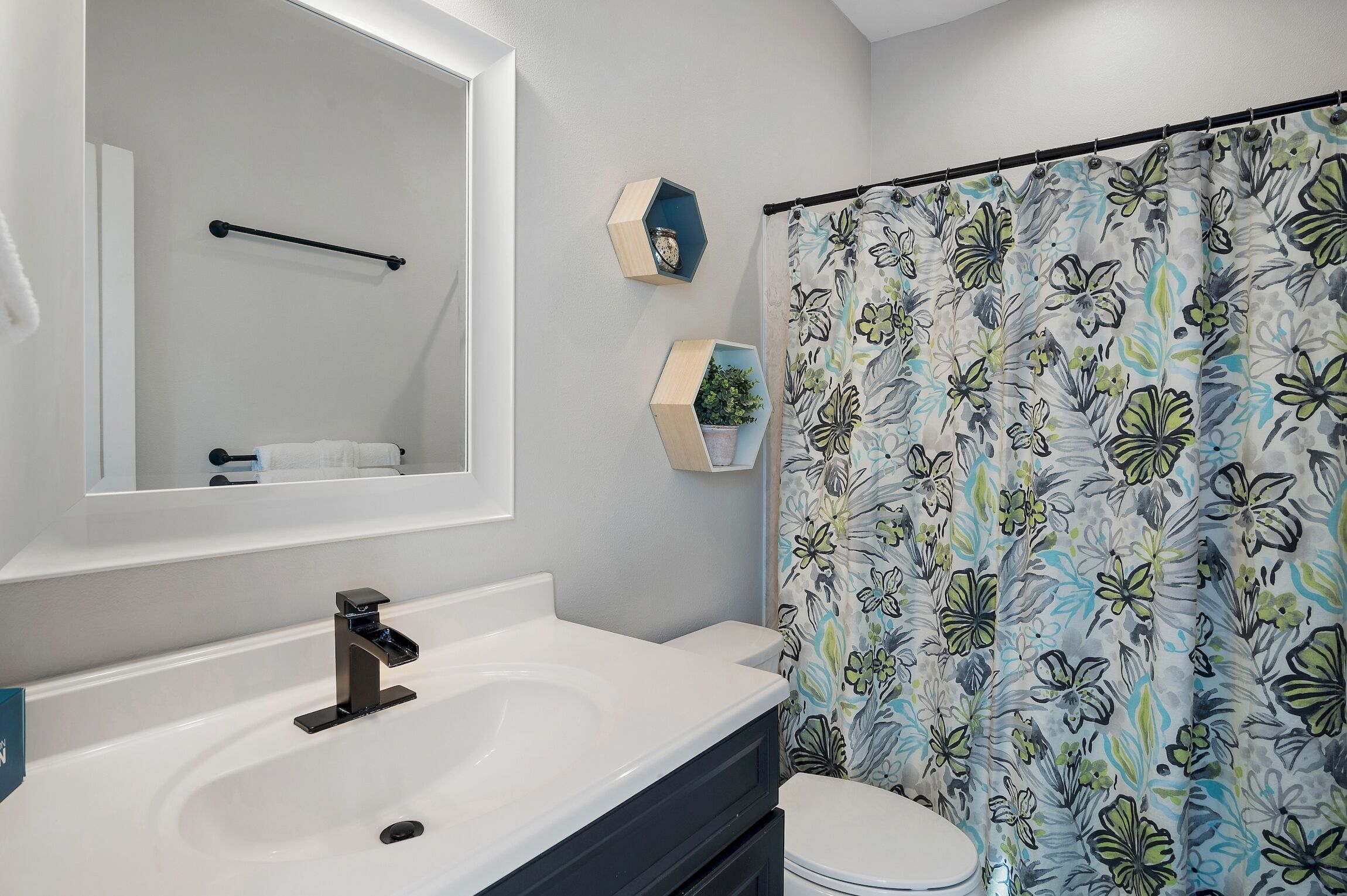 A modern bathroom with a white sink, black faucet, and a toilet. A shower curtain with a floral pattern is visible.
