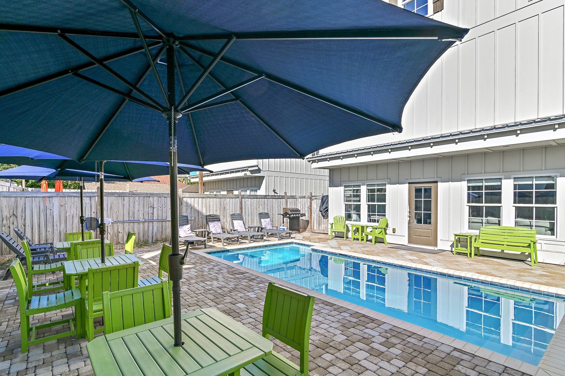 A swimming pool with green chairs and umbrellas, surrounded by a wooden fence and a white building.
