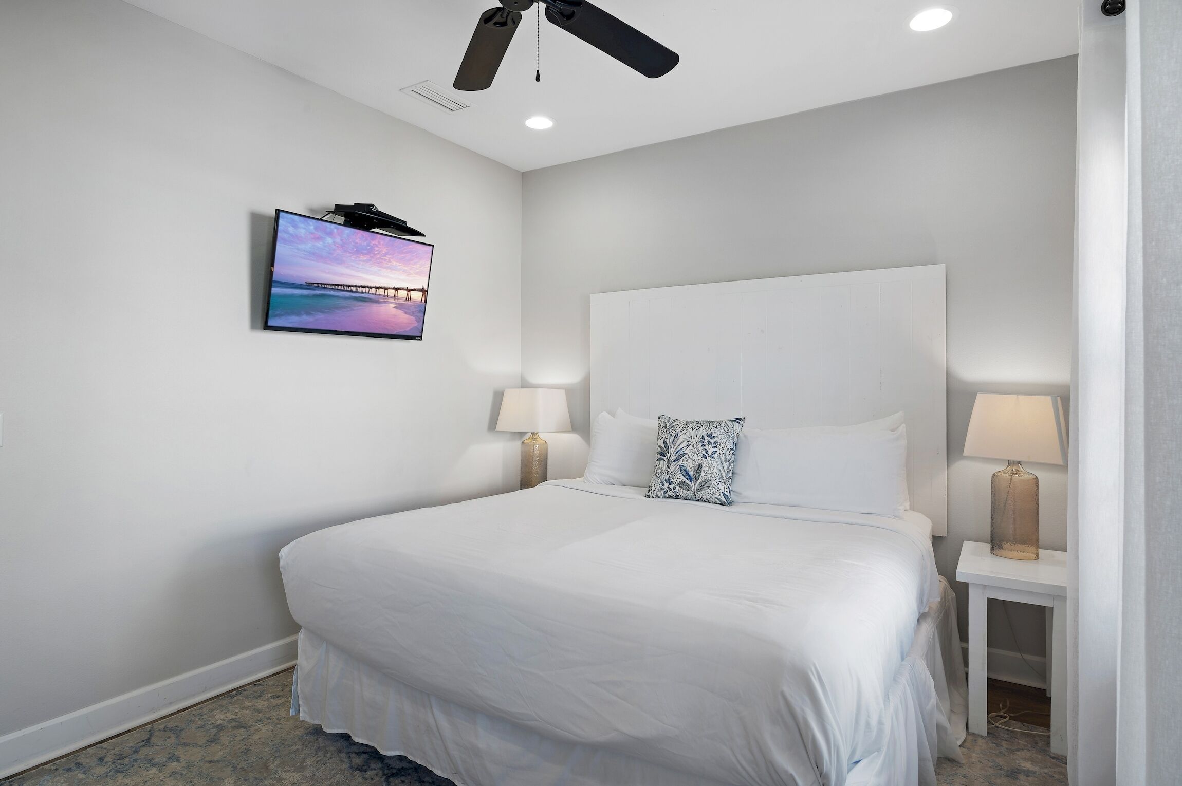 A modern hotel room with a large bed, two bedside lamps, a ceiling fan, and a TV mounted on the wall.