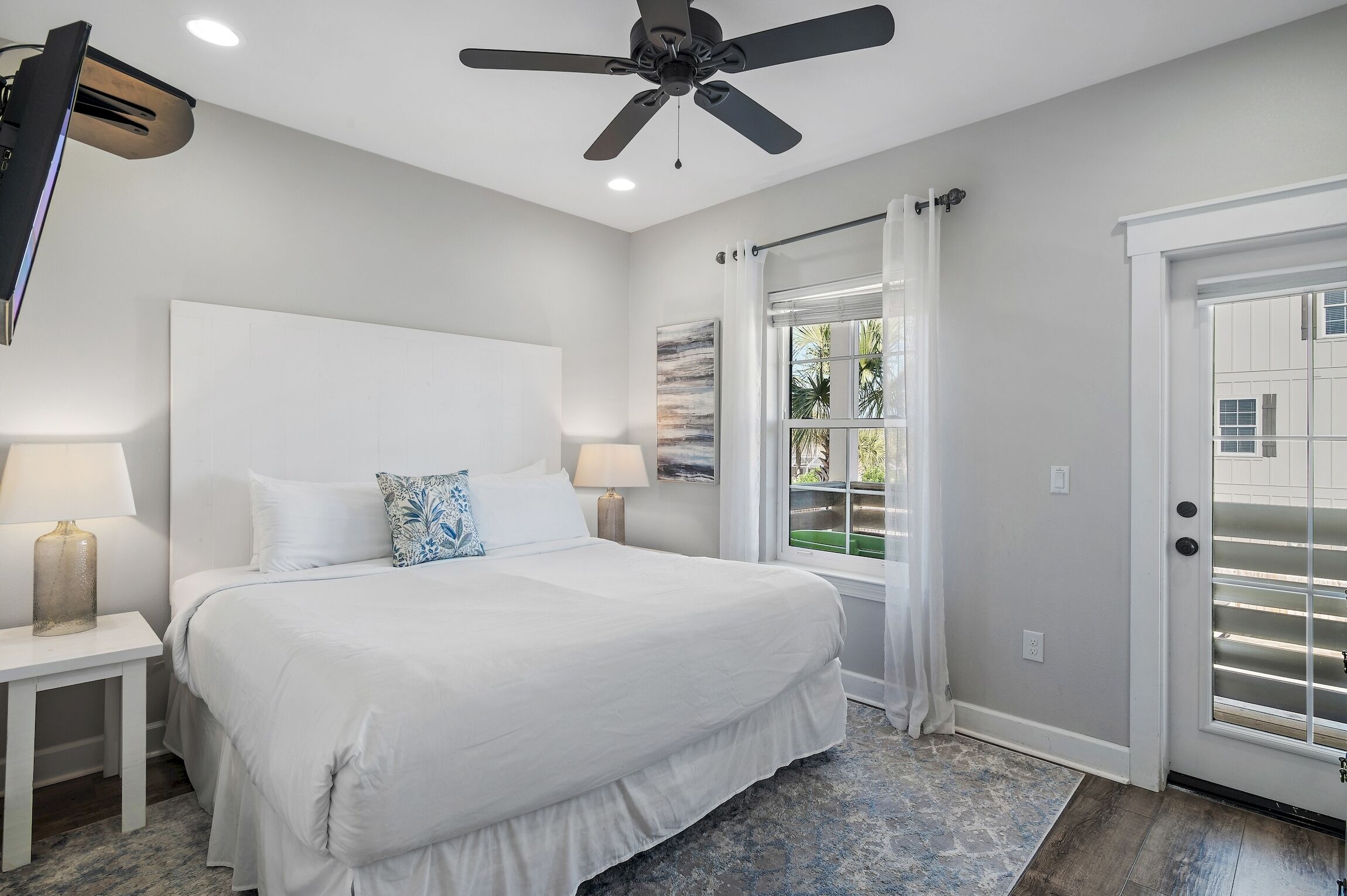 A bedroom with a large bed, a ceiling fan, a window with curtains, and a door with blinds.