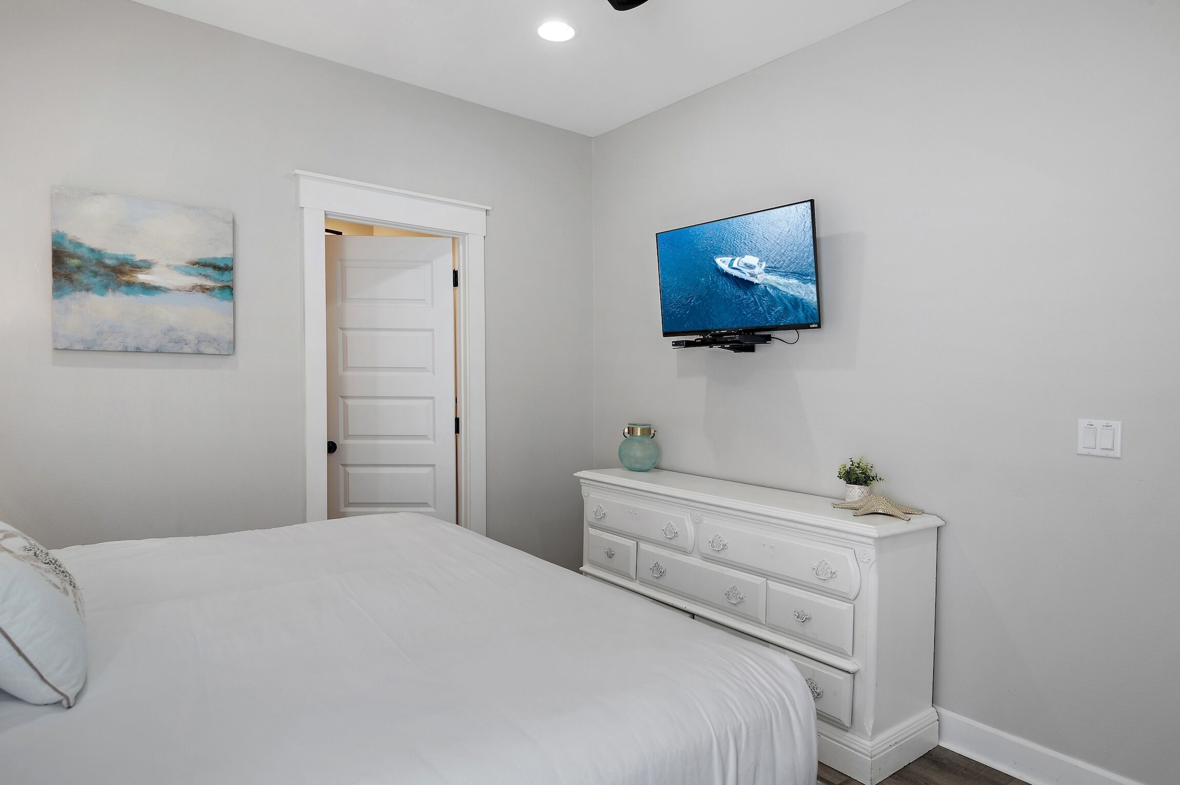 Une chambre à coucher comprenant un lit, une commode, un téléviseur fixé au mur et une porte donnant sur une autre pièce.
