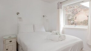 Une chambre à coucher blanche, avec un lit, une table de chevet, une fenêtre et une vue sur un mur de briques à l’extérieur.