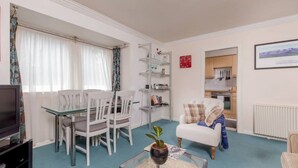 Un salon avec une table basse en verre, un canapé blanc et un téléviseur. On y trouve également un coin repas avec une table et des chaises. Des étagères remplies de livres et d’objets décoratifs sont visibles.