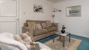 Un salon avec un canapé beige, un fauteuil blanc, une table basse en verre, un tapis à motifs, une plante en pot, un tableau accroché au mur, un lampadaire et un tableau encadré.