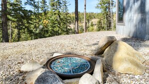 A metal fire pit with a blue mosaic design surrounded by rocks and pebbles, set outdoors with trees and a building in the background.