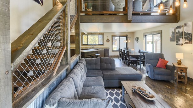 A spacious living area with a large gray sectional sofa, a wooden coffee table, and a dining area with a table and chairs. A staircase with a wooden railing leads to an upper level.