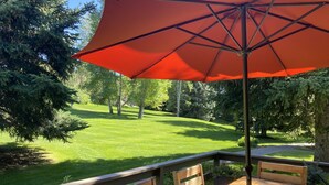 A wooden deck with a table and chairs, an orange umbrella, and a view of a well-maintained lawn and trees. - Modern rustic condo in the heart of iconic Sun Valley (Sun Valley)