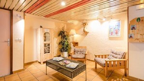 A cozy room with a wooden ceiling, a small table with magazines, a chair, a lamp, and a plant.