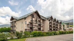 A multi-story hotel with balconies, a parking area, and a mountainous backdrop.