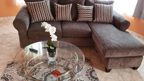 En modulsofa med mønstrede hynder, et sofabord i glas med en fjernbetjening og en potteplante, samt en tæppe med et mønster på et flisebelagt gulv.