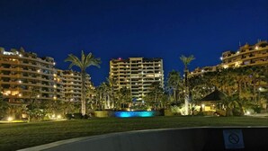 Vista nocturna de un complejo turístico con varios edificios altos, una piscina y palmeras.