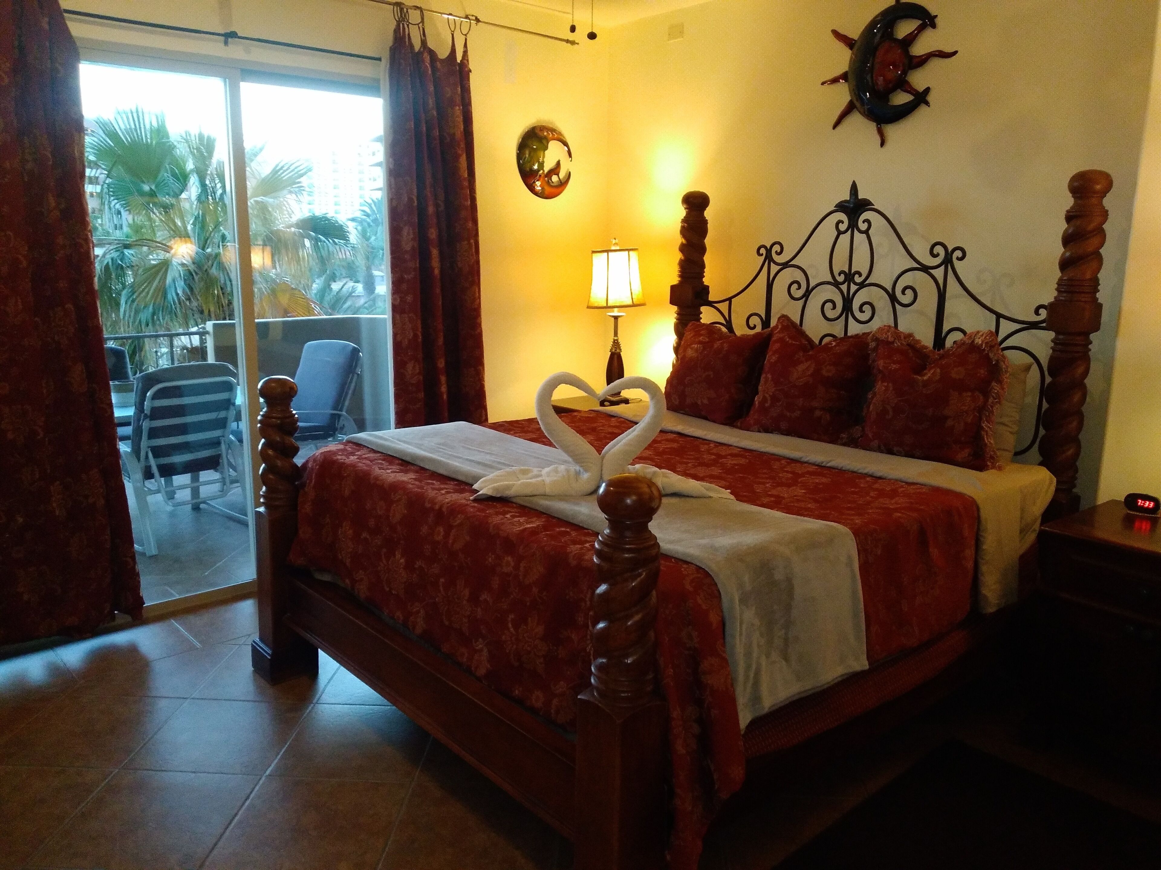 A hotel room with a bed, a nightstand, a lamp, a clock, and a view of palm trees through the window.