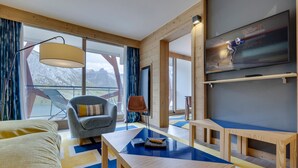 Une chambre d’hôtel moderne dotée d’une grande fenêtre offrant une vue sur les montagnes, d’un canapé confortable, d’une table basse en bois et d’un téléviseur à écran plat fixé au mur.