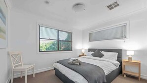 A bedroom with a bed, two bedside tables, a chair, and two windows with blinds. - Noble House~2 Bed/1Bath/1Car~Parklands + Transport (Wilston)