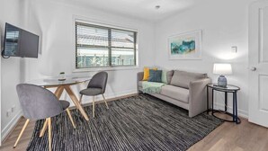 A modern living room with a sofa, a TV, a table, and a window with blinds. - Noble House~2 Bed/1Bath/1Car~Parklands + Transport (Wilston)