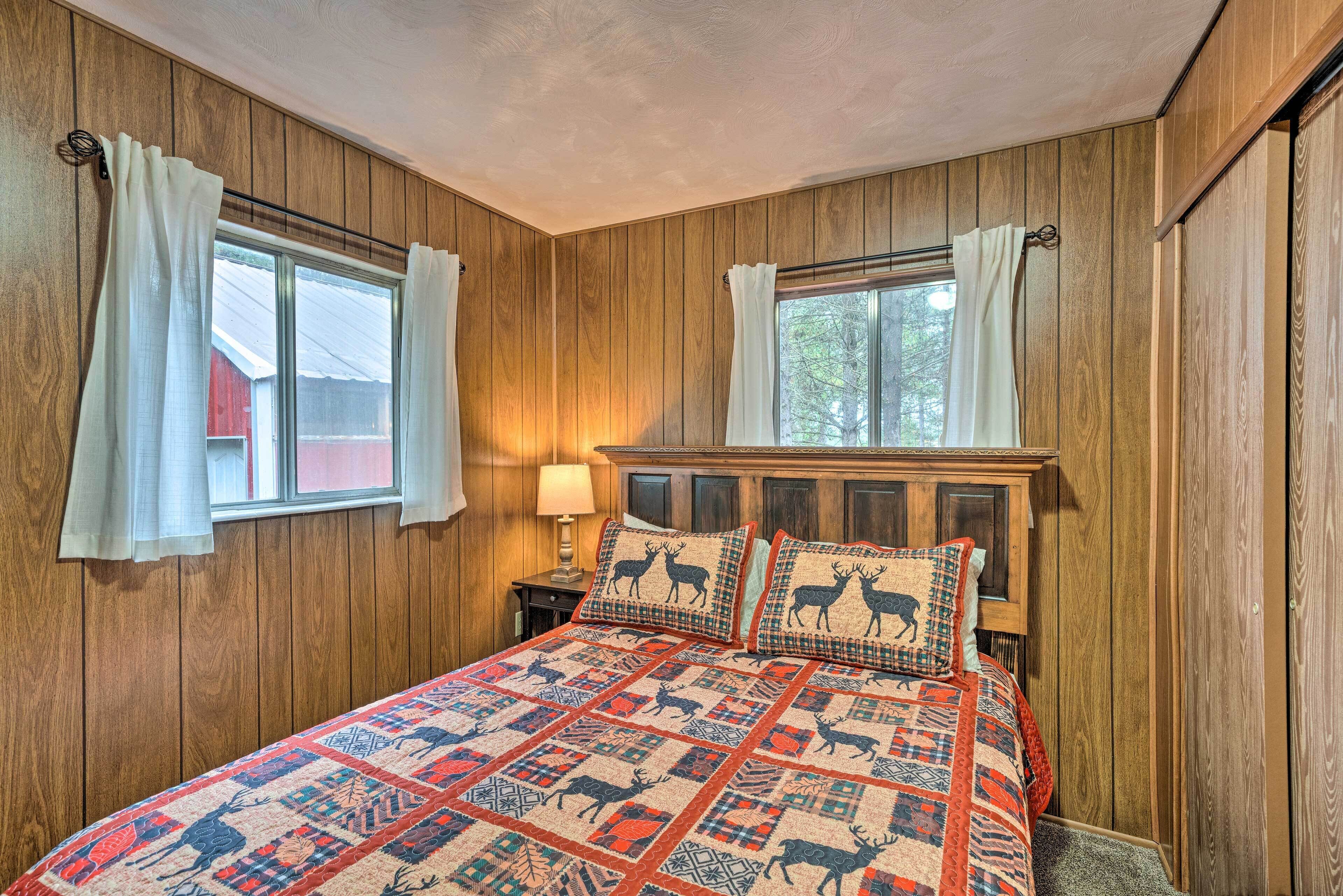 Un dormitorio con paredes de paneles de madera, una cama con colcha estampada, dos ventanas con cortinas y una mesita de noche con lámpara.