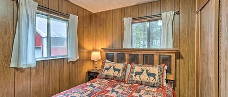 Un dormitorio con paredes de paneles de madera, una cama con colcha estampada, dos ventanas con cortinas y una mesita de noche con lámpara.