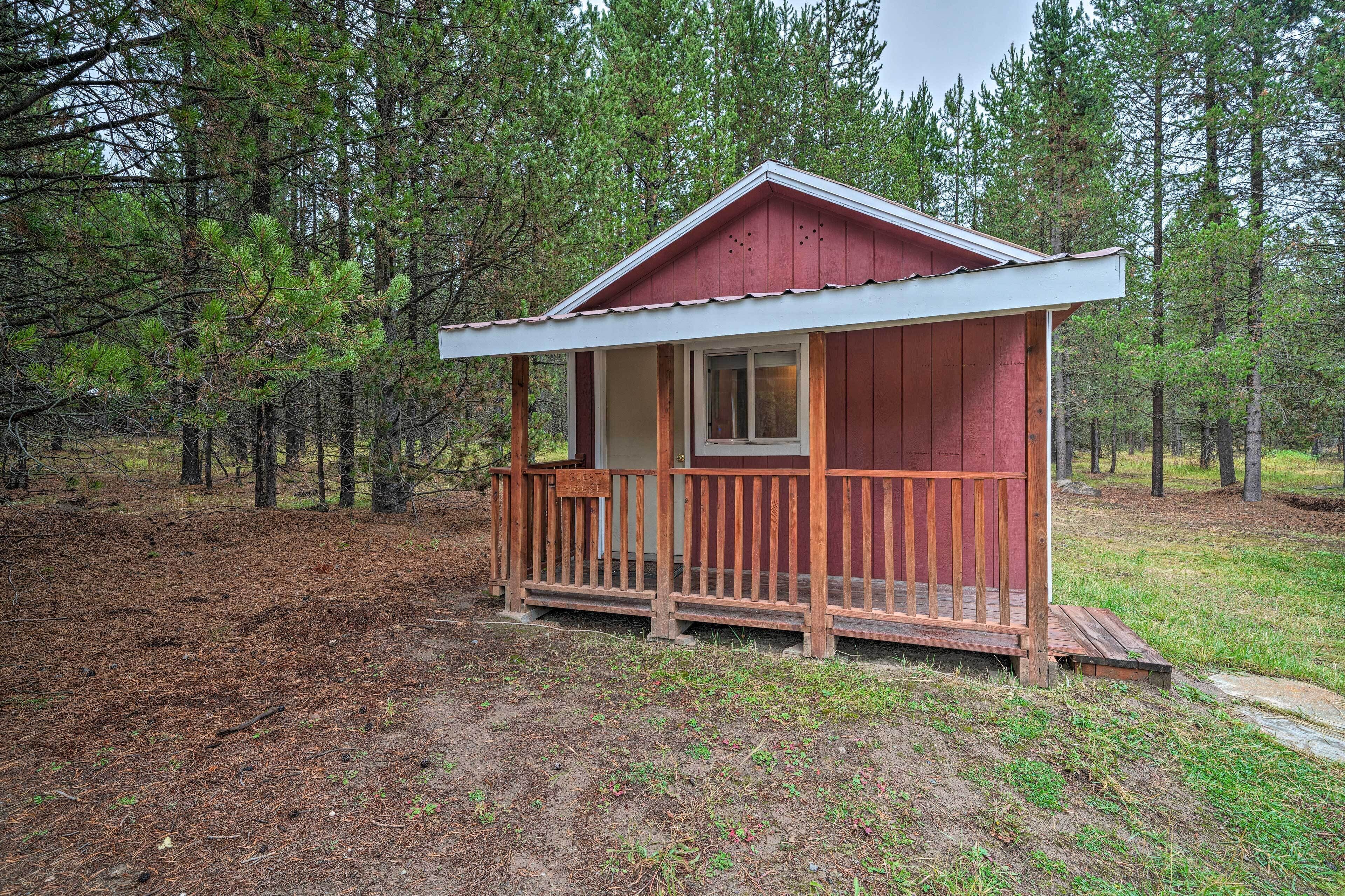A small cabin with a wooden deck and a single window, surrounded by a forest.