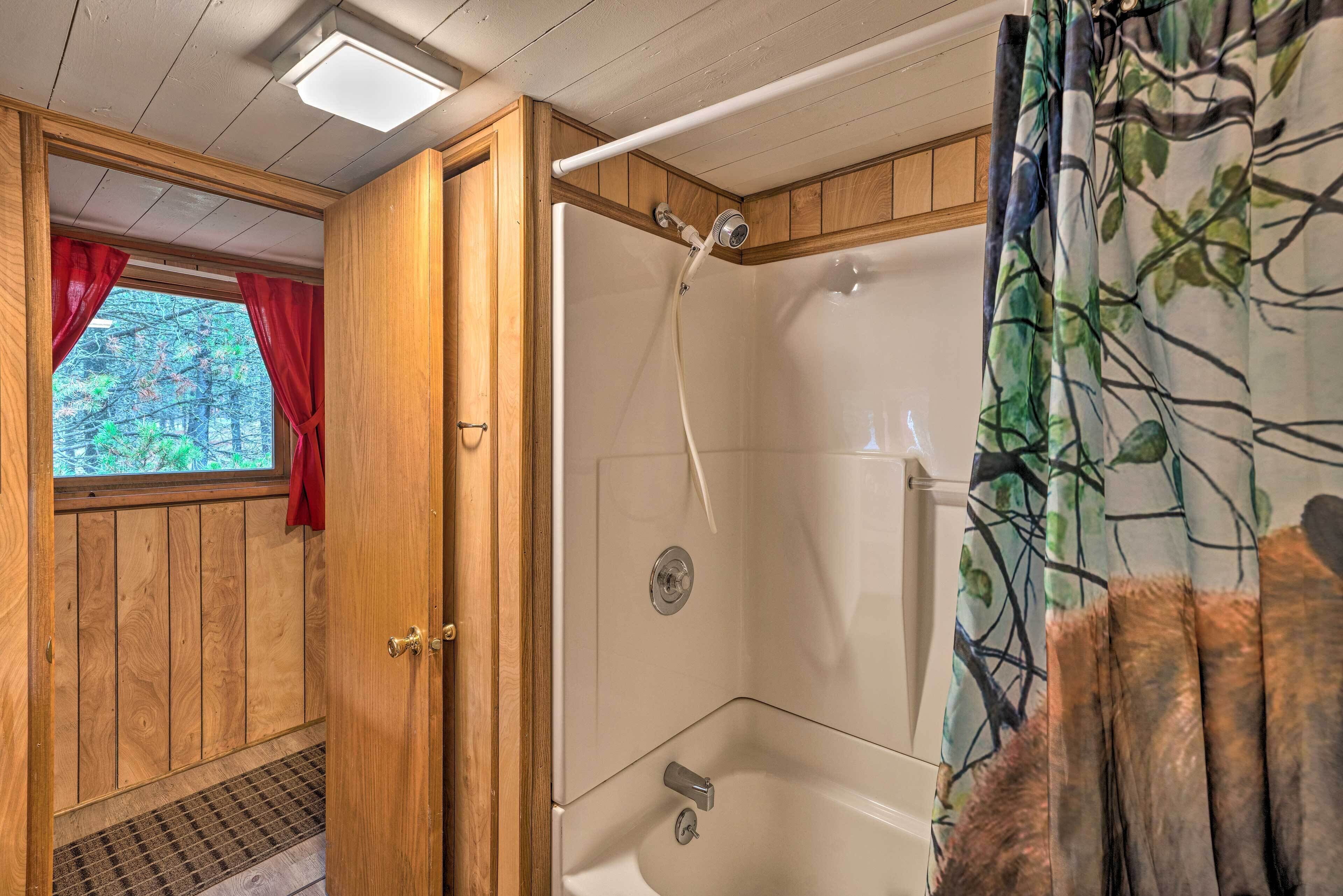 A wooden cabin interior with a shower area, a window with red curtains, and a view of trees.