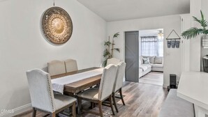 A dining area with a wooden table and upholstered chairs, a decorative wall piece, and a view into a living room with a sofa and plants. - Beautiful house to relax and enjoy close to Camarillo outlets and beach (Camarillo)